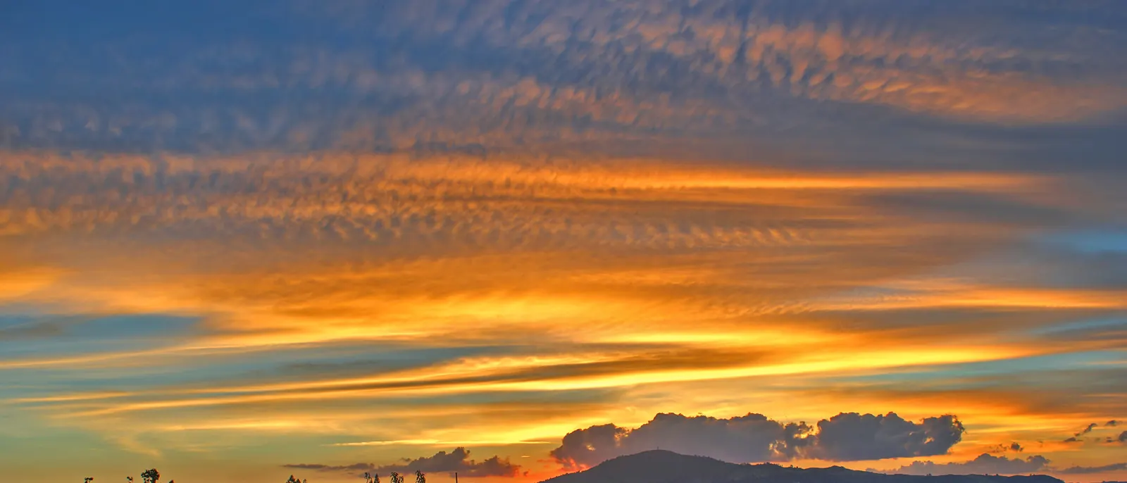 A sunset sky of orange, gold and blue bands of cloud over the rooftops of Lynmore, Rotorua, with a low hill on the horizon